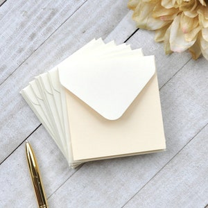 May include: A stack of ivory envelopes with a white inner flap. The envelopes are arranged in a fan shape on a light wood surface.