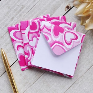 May include: A stack of pink and white patterned envelopes with a white interior. The envelopes feature a repeating pattern of pink hearts.