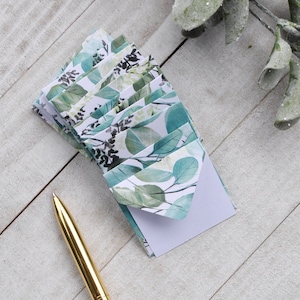 May include: A stack of small, white envelopes with a green and white floral pattern. The envelopes are on a white wooden surface with a gold pen.
