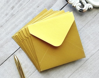 May include: A stack of gold envelopes with a yellow flap. The envelopes are arranged in a fan shape and are lying on a light wood surface.