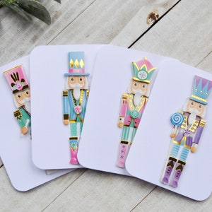May include: Four decorative Christmas cards featuring colorful nutcracker soldier designs. Each card showcases a different nutcracker with varying color schemes, including pink, blue, and purple, with gold accents. A purple pen rests on the right.