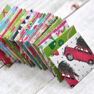 May include: A collection of colorful, folded paper squares with various Christmas-themed patterns. Designs include red cars carrying Christmas trees, candy canes, and geometric shapes in green, red, and blue. The papers are arranged on a white wooden surface.