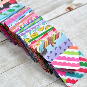 May include: A stack of colorful patterned mini envelopes. The envelopes are folded and stacked on a white wooden surface. The top envelope has a pink and white striped pattern with a scalloped edge.
