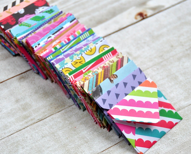 May include: A stack of colorful patterned mini envelopes. The envelopes are folded and stacked on a white wooden surface. The top envelope has a pink and white striped pattern with a scalloped edge.