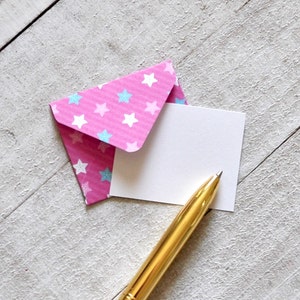 May include: A pink envelope with blue and white stars printed on it. A blank white card is inside the envelope. A gold pen is lying on top of the card.