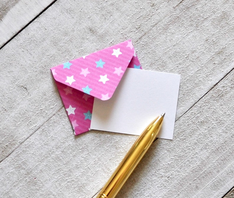 May include: A pink envelope with blue and white stars printed on it. A blank white card is inside the envelope. A gold pen is lying on top of the card.