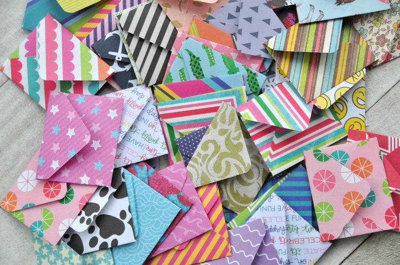 May include: A collection of colorful patterned paper envelopes, some with stars, polka dots, stripes, and other designs. The envelopes are arranged in a pile on a white surface.