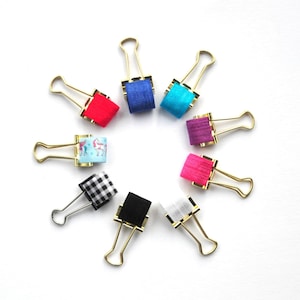 May include: A set of ten gold binder clips with colorful patterned ribbon accents. The clips are arranged in a circle on a white background.