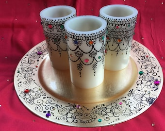 Puede incluir: Tres velas blancas con intrincados diseños de henna negra y brillantes de colores se muestran en un plato dorado con un diseño de henna negra.