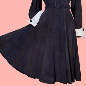 May include: A dark navy blue dress with a flared skirt and long sleeves. The dress features white cuffs with black buttons and a matching belt. The fabric appears to be suede or a similar material, with a flowing design.