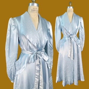 May include: A light blue satin robe featuring a shawl collar, long sleeves, and a tie waist. The garment is accented with textured, quilted-style panels on the collar, cuffs, pockets, and hemline, displayed on two dress forms against a gold background.