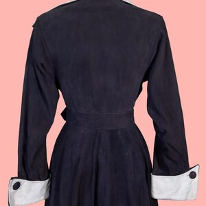 May include: A navy blue vintage coat with a light blue collar and white cuffs. The coat has a belted waist and long sleeves. The cuffs feature a black button detail. The coat is displayed on a mannequin against a pink background.