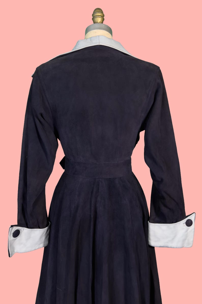 May include: A navy blue vintage coat with a light blue collar and white cuffs. The coat has a belted waist and long sleeves. The cuffs feature a black button detail. The coat is displayed on a mannequin against a pink background.