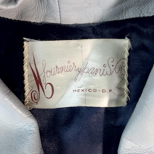 May include: Close-up of a light gray leather jacket with a dark blue lining. The jacket's label reads "Fournier Paris S.A. MEXICO D.F." in a cursive font. The jacket appears to be vintage.