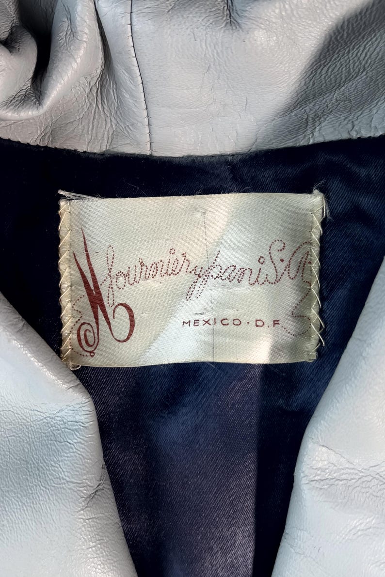May include: Close-up of a light gray leather jacket with a dark blue lining. The jacket's label reads "Fournier Paris S.A. MEXICO D.F." in a cursive font. The jacket appears to be vintage.