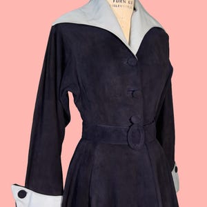 May include: A vintage navy blue dress with a light gray collar and cuffs. The dress features long sleeves, a belted waist, and button details. The dress is displayed on a mannequin against a pink background.