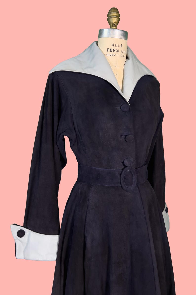 May include: A vintage navy blue dress with a light gray collar and cuffs. The dress features long sleeves, a belted waist, and button details. The dress is displayed on a mannequin against a pink background.