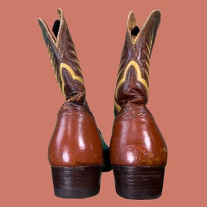 May include: A pair of brown and yellow cowboy boots. The boots have a pointed toe and a tall shaft with decorative stitching. The heels are dark brown, and the boots appear to be made of leather. The background is a solid peach color.