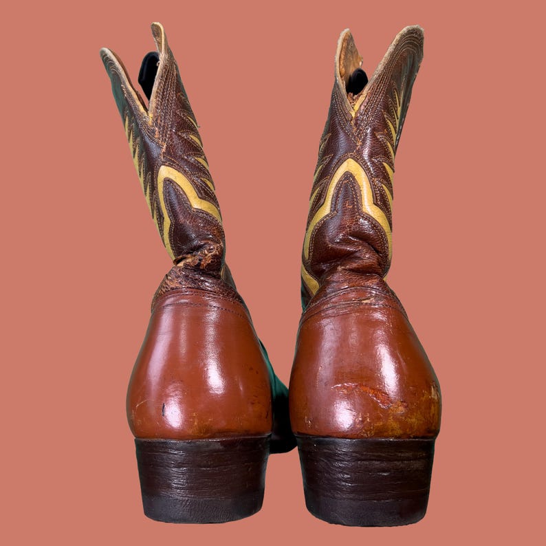 May include: A pair of brown and yellow cowboy boots. The boots have a pointed toe and a tall shaft with decorative stitching. The heels are dark brown, and the boots appear to be made of leather. The background is a solid peach color.