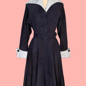 May include: A vintage navy blue coat with a light gray collar and cuffs. The coat features a belted waist, long sleeves, and button closures. The coat is displayed on a mannequin against a pink background.