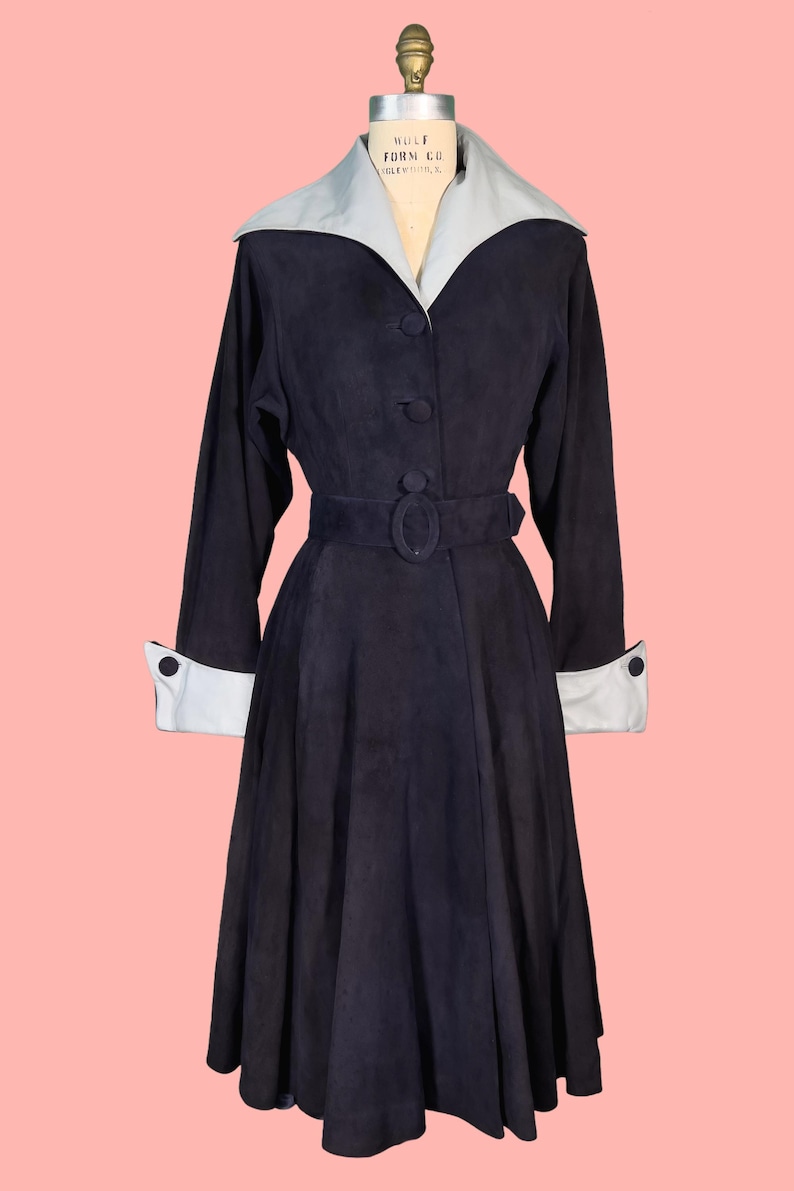 May include: A vintage navy blue coat with a light gray collar and cuffs. The coat features a belted waist, long sleeves, and button closures. The coat is displayed on a mannequin against a pink background.