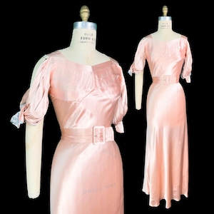 Könnte beinhalten: Zwei Vintage-Kleider aus hellrosa Satin, die auf Schneiderpuppen präsentiert werden. Die Kleider haben einen quadratischen Ausschnitt, kurze Ärmel und einen passenden Gürtel mit quadratischer Schnalle. Der Stoff hat einen dezenten Glanz und die Kleider haben eine klassische, elegante Silhouette.
