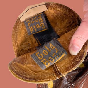 May include: Close-up of a brown leather cowboy boot. The boot's interior reveals a label that reads "Gold Bond" in yellow lettering on a gray background. The boot has a worn, textured appearance.