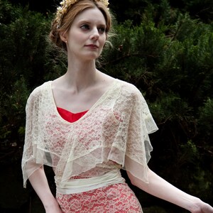 May include: A woman wearing a cream-colored lace dress with a red underdress. The dress features a V-neckline and a flowing skirt. A cream-colored lace shawl is draped over the shoulders. A floral crown adorns the woman's hair.