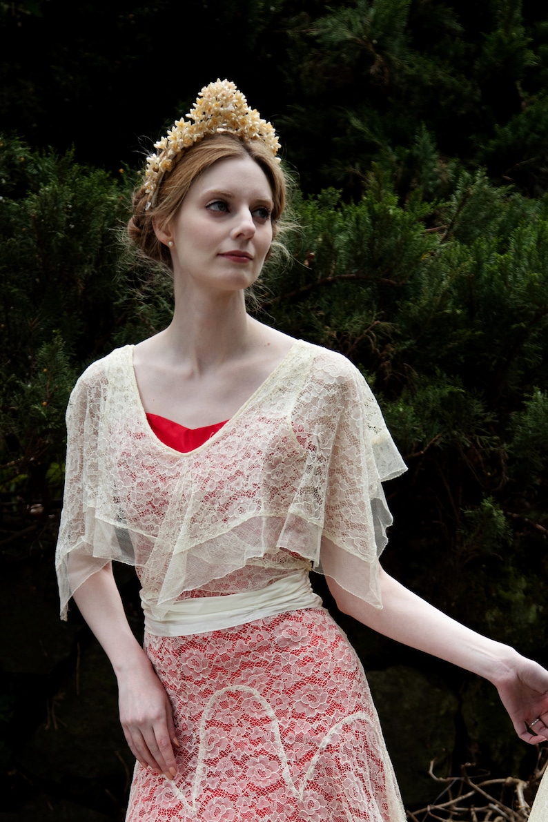 May include: A woman wearing a cream-colored lace dress with a red underdress. The dress features a V-neckline and a flowing skirt. A cream-colored lace shawl is draped over the shoulders. A floral crown adorns the woman's hair.