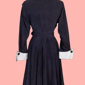 May include: A navy blue vintage coat with a light blue collar and white cuffs. The coat features a belted waist and button details on the cuffs. The coat is displayed on a mannequin against a pink background.