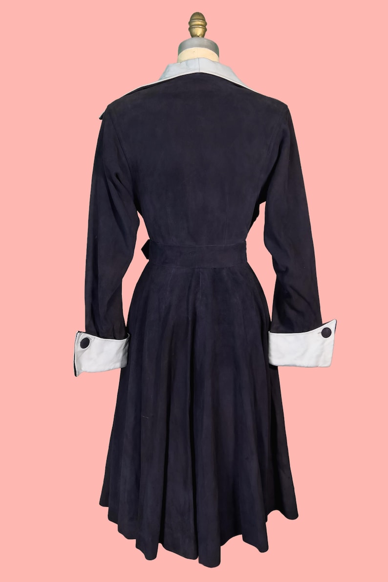 May include: A navy blue vintage coat with a light blue collar and white cuffs. The coat features a belted waist and button details on the cuffs. The coat is displayed on a mannequin against a pink background.