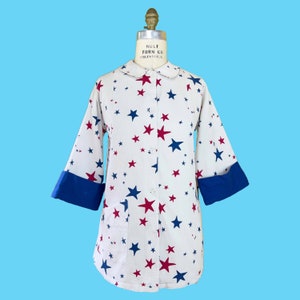 May include: A white button-up shirt with a blue and red star pattern. The shirt has a collar and long sleeves with blue cuffs. The shirt is on a mannequin.