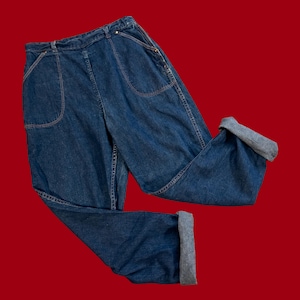 Peut inclure: Jean bleu avec de grandes poches avant et des revers aux chevilles. Le jean a des coutures apparentes de couleur contrastante et une coupe décontractée. Les revers sont gris. Le jean est présenté sur fond rouge.