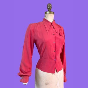 May include: A coral-coloured vintage blouse with long sleeves and a pointed collar. The blouse features a single chest pocket and button closures down the front and on the cuffs. The garment is displayed on a mannequin against a purple background.