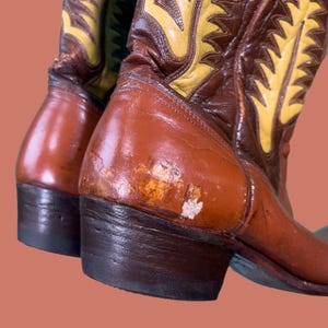 May include: Close-up of a pair of brown and yellow cowboy boots. The boots have a distressed appearance with visible wear and tear on the leather. The design includes decorative stitching and a classic western style.