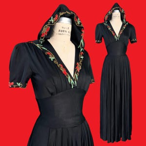 May include: A black floor-length dress featuring a hood, short sleeves, and a deep V-neckline. The hood and neckline are embellished with red and green floral embroidery. The dress has a fitted waist and a pleated skirt, displayed on a mannequin.