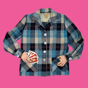 May include: A long-sleeved button-down jacket featuring a plaid pattern in shades of blue, black, and cream. The jacket includes a pointed collar, two front patch pockets, and white buttons. A hand holds a small fan of playing cards over the pocket.