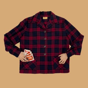 May include: A long-sleeved button-down shirt featuring a dark navy blue and deep red plaid pattern. The shirt includes a pointed collar, four dark buttons down the center front, and two square patch pockets at the lower front hem area.