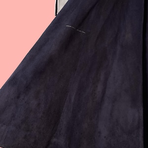 May include: A dark navy blue suede coat with white trim and black polka dots on the sleeves. The coat has a flared skirt and a soft, textured appearance. The background is a light pink.