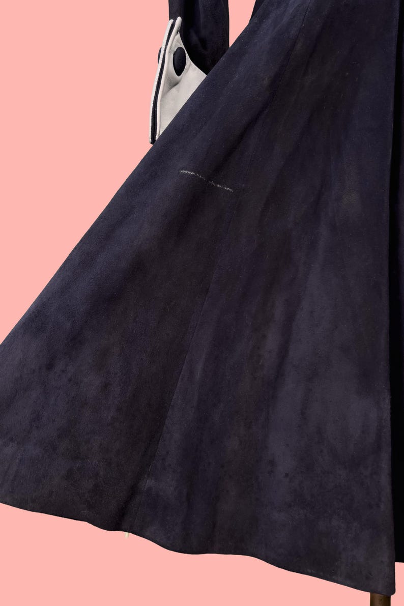 May include: A dark navy blue suede coat with white trim and black polka dots on the sleeves. The coat has a flared skirt and a soft, textured appearance. The background is a light pink.