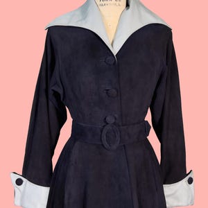 May include: A vintage navy blue coat with a light gray collar and cuffs. The coat features a belted waist, button closures, and long sleeves. The coat is displayed on a mannequin against a pink background.
