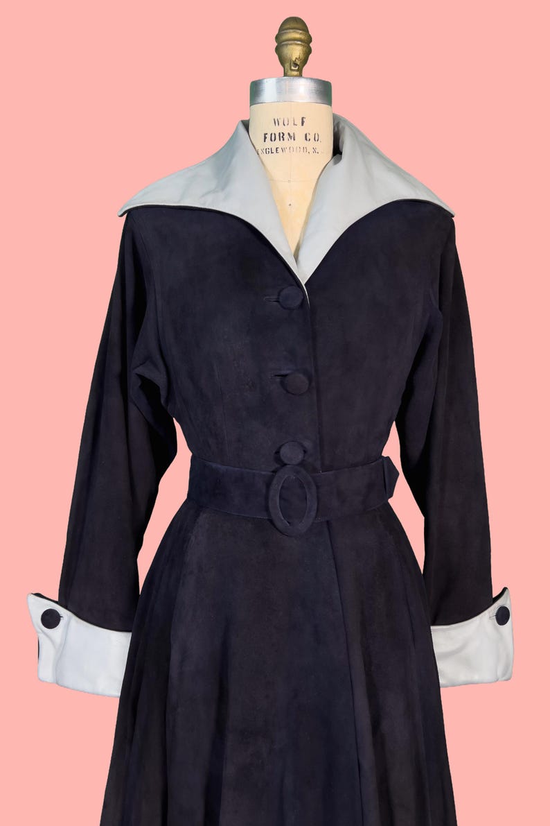 May include: A vintage navy blue coat with a light gray collar and cuffs. The coat features a belted waist, button closures, and long sleeves. The coat is displayed on a mannequin against a pink background.