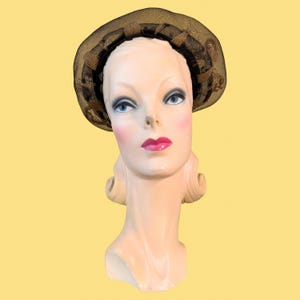 Pode incluir: Um chapéu vintage com uma copa de malha castanha escura e uma faixa decorativa. O chapéu é exibido em uma cabeça de manequim com olhos azuis, bochechas rosadas e batom vermelho. A aba do chapéu é adornada com detalhes castanhos.