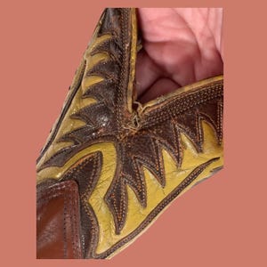 May include: Close-up of a vintage cowboy boot. The boot features a brown and yellow leather design with intricate stitching. The toe and heel are a darker brown, while the shaft has a lighter yellow color with decorative patterns.