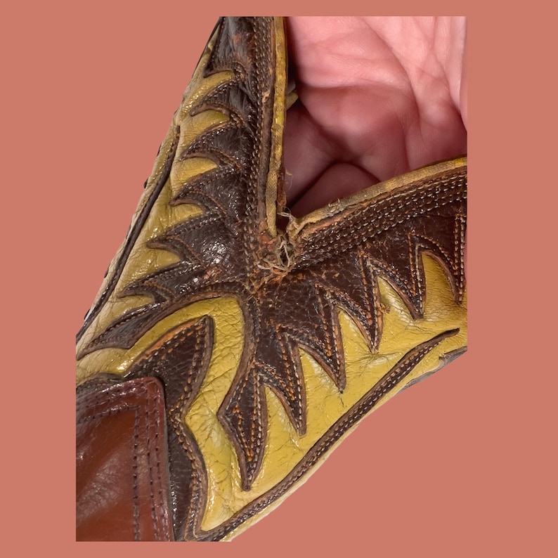 May include: Close-up of a vintage cowboy boot. The boot features a brown and yellow leather design with intricate stitching. The toe and heel are a darker brown, while the shaft has a lighter yellow color with decorative patterns.