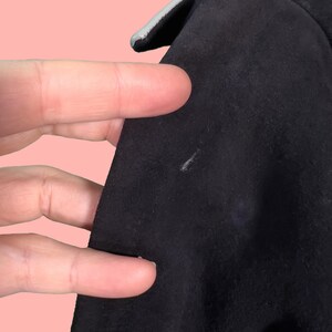 May include: Close-up of a black garment with a light gray trim. The fabric appears to be a soft, suede-like material. The image shows a portion of the garment's edge and a small area of the fabric's surface.