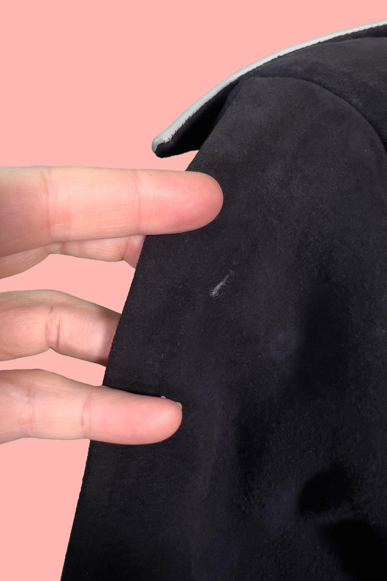 May include: Close-up of a black garment with a light gray trim. The fabric appears to be a soft, suede-like material. The image shows a portion of the garment's edge and a small area of the fabric's surface.
