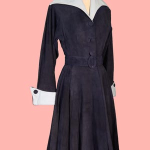 May include: A vintage navy blue dress with a light blue collar and cuffs. The dress features long sleeves, a belted waist, and a button-down front. The dress is displayed on a mannequin against a pink background.
