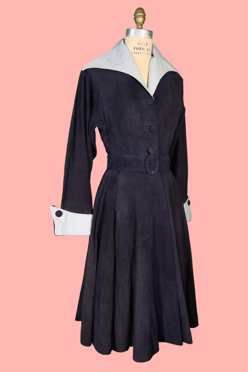 May include: A vintage navy blue dress with a light blue collar and cuffs. The dress features long sleeves, a belted waist, and a button-down front. The dress is displayed on a mannequin against a pink background.