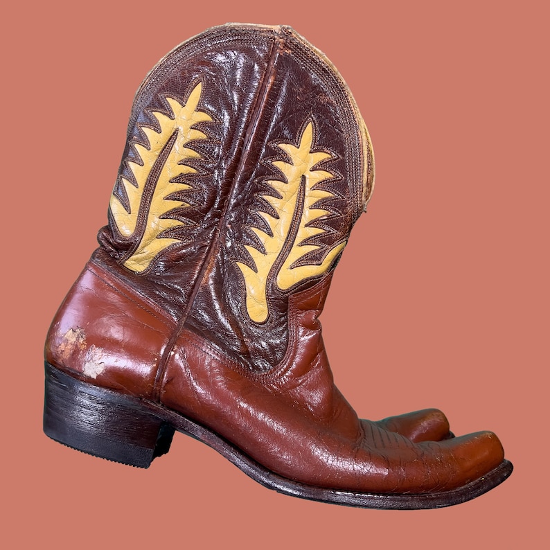 May include: Brown leather cowboy boot with a pointed toe and a decorative yellow and brown stitched design on the shaft. The boot has a worn appearance, with scuffs and marks on the leather.